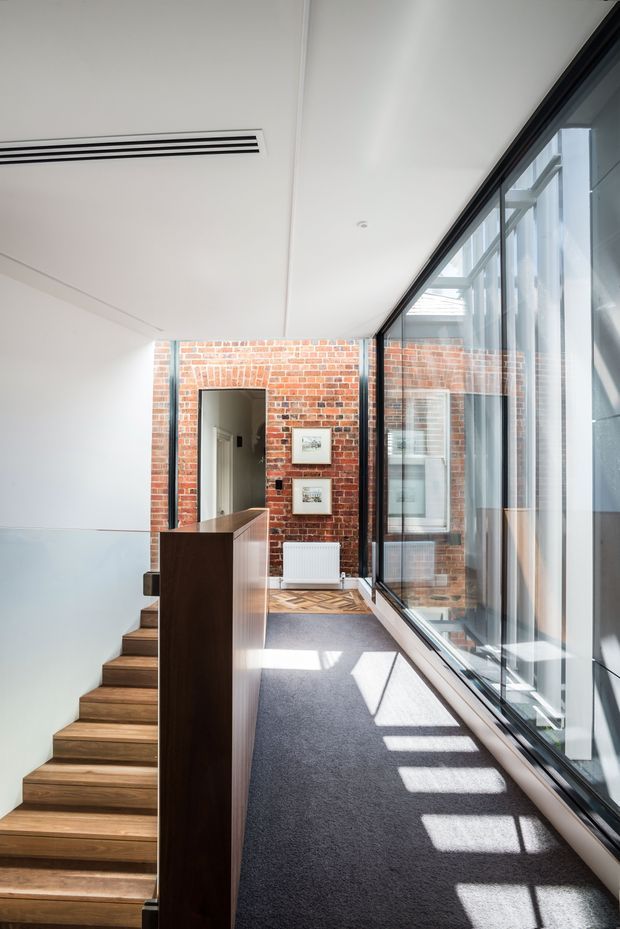 A bright, narrow interior landing beside a staircase. The space features a dark grey carpeted floor, warm timber stair treads and balustrade, a white ceiling with linear air-conditioning vents, exposed red brick walls, and extensive black-framed glazing along one side. Strong sunlight casts geometric shadows across the carpet.