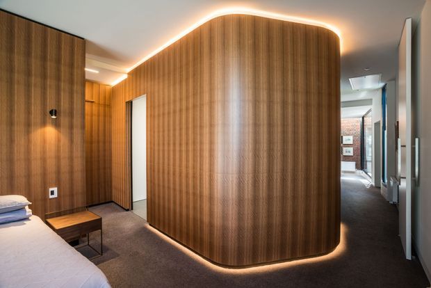 Contemporary bedroom interior featuring a dramatic curved, floor-to-ceiling timber-panelled partition or built-in wardrobe with concealed warm LED lighting around its edges. The room has dark carpet, white bedding, a compact timber bedside table, and a hallway or adjoining space visible beyond the partition.
