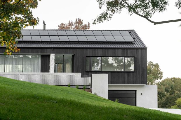 Exterior view of a contemporary dark-clad house on a sloping, landscaped site. The single-storey-looking form has a pitched metal roof covered with solar panels, large horizontal windows, pale masonry or rendered wall sections, and an integrated garage partly set below the house. Trees and lawn surround the building.