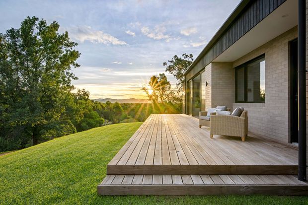 A modern single-storey home viewed from a broad timber deck at sunset. The exterior features pale brick walls, dark vertical cladding along the upper facade, expansive glazing, deep roof overhangs, and a built-in outdoor seating area. The deck steps down toward a manicured lawn, with a leafy landscape and distant sunset beyond.