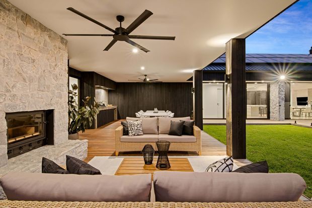 A refined indoor-outdoor living space with a large sectional sofa facing a stone fireplace, dark timber feature walls, timber flooring and a dining area beyond. Full-height openings connect the living room to a landscaped courtyard lawn and a covered outdoor entertaining area. A large ceiling fan, recessed lighting and exposed dark structural posts contribute to the contemporary pavilion-like design.
