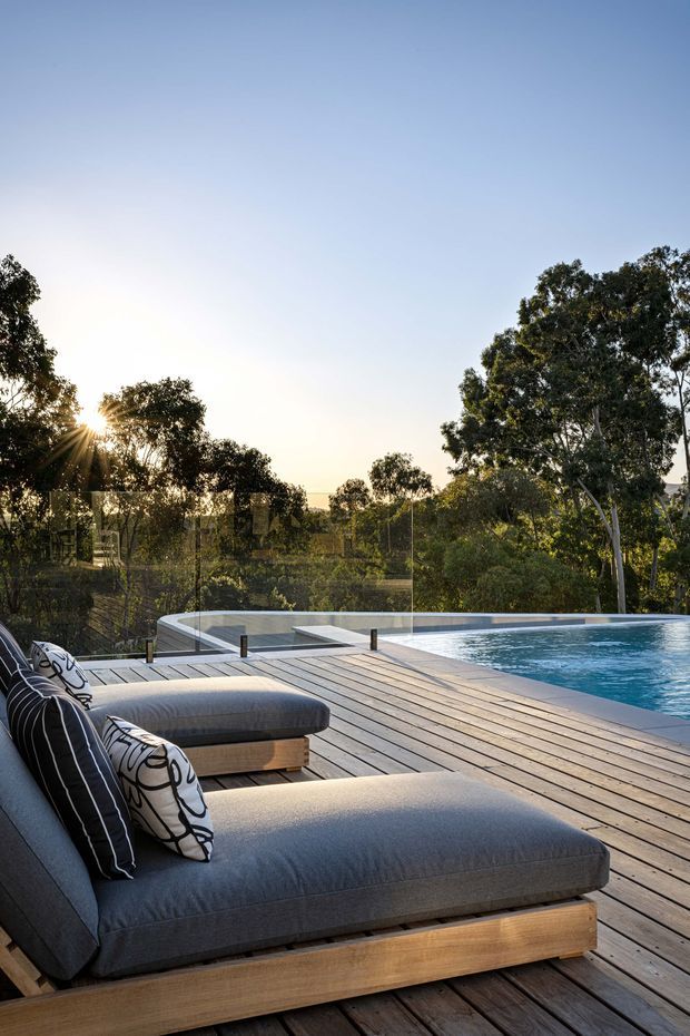 A serene outdoor pool deck at sunset, with large grey upholstered timber-framed sun loungers in the foreground, a broad timber decking platform, glass pool fencing, and a rectangular swimming pool overlooking a leafy landscaped setting.