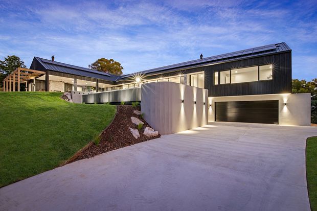 A dusk exterior view of a contemporary single-storey home on a gently sloping landscaped site. The house has dark vertical cladding, long low-pitched roof forms with solar panels, extensive glazed openings, a covered outdoor entertaining area, and an integrated double garage. A broad concrete driveway and illuminated retaining wall lead toward the entry, with a manicured lawn in the foreground.