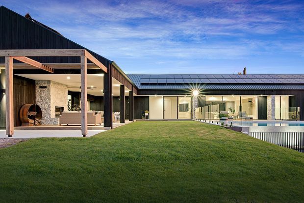 Exterior view of a contemporary single-storey residence arranged around a broad lawn and pool terrace. The home features dark vertical cladding, low-pitched metal roofs, extensive glazing, timber-framed covered outdoor living, and a stone-clad fireplace wall. A pool and recessed outdoor area are visible on the right.
