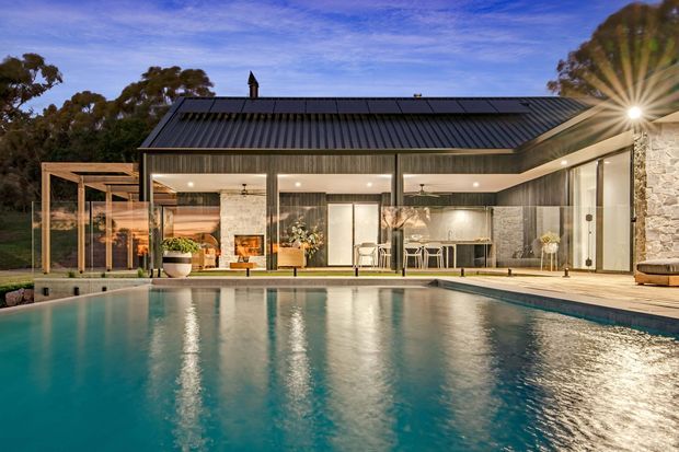 Contemporary single-storey residence viewed from a swimming pool at dusk. The home features a dark metal pitched roof, extensive floor-to-ceiling glazing, vertical timber wall cladding, pale stone walls, and a covered outdoor entertaining area with a kitchen or bar opening onto the pool terrace. Mature trees surround the property.