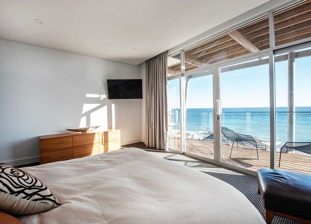Bright contemporary bedroom with a large bed, low timber dresser, wall-mounted television, and full-height glazed doors opening onto a timber deck overlooking the ocean. The room has a white minimalist fit-out, soft curtains, and strong natural light, with a partial outdoor seating area visible beyond the glazing.