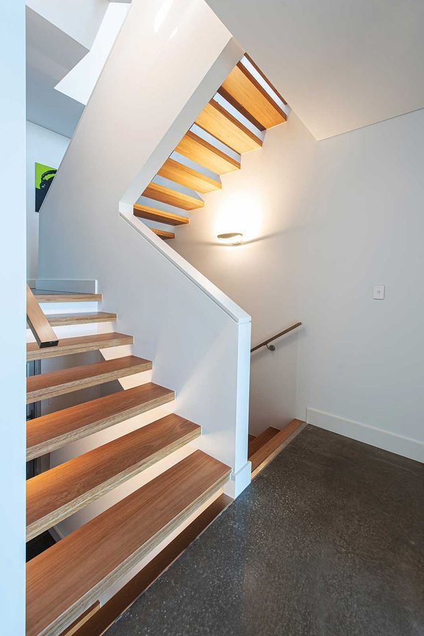 Bright modern stairway interior with open-riser timber stairs, white painted walls and balustrade, and a second flight visible above. The stairwell has a dark polished floor, timber handrail, recessed wall lighting, and strong natural light from above.