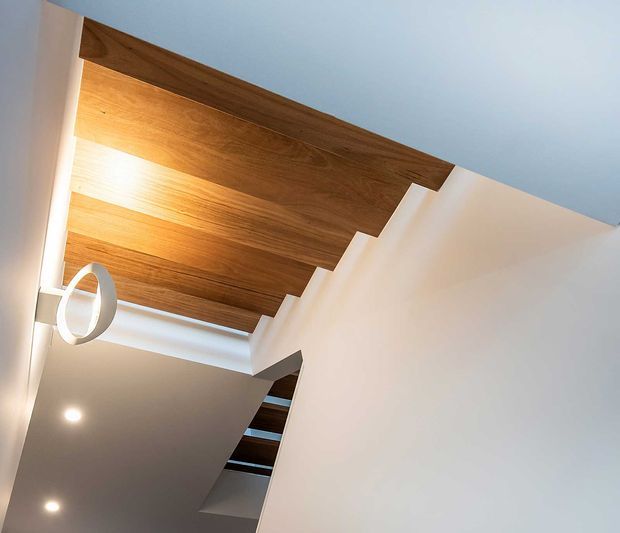 Interior architectural detail showing a contemporary stairway with white walls and a warm timber-lined ceiling or soffit. Dark stair treads and open riser gaps are visible, along with recessed downlights and a circular pendant light. The image is rotated, emphasizing the ceiling planes, stair geometry, and clean minimalist detailing.