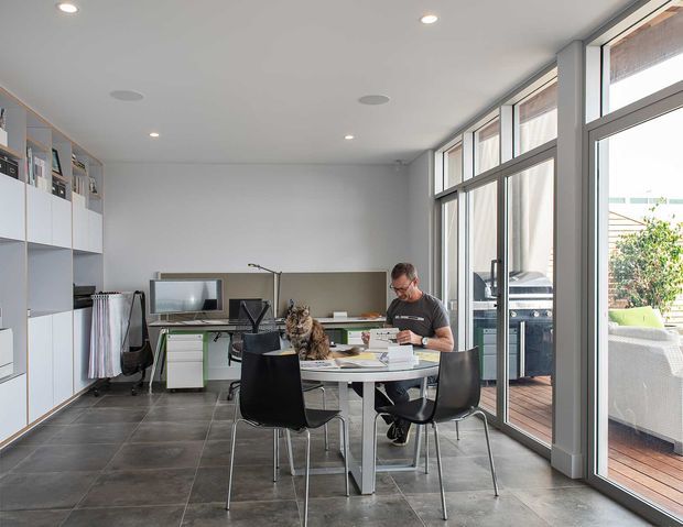 Bright contemporary home office and dining workspace with a large rectangular table, black chairs, computer workstation, built-in white shelving and cabinetry, and full-height glazed doors and windows opening to a timber deck. The room has a simple white ceiling with recessed and round acoustic-style fittings, dark large-format tiled flooring, and abundant natural light.