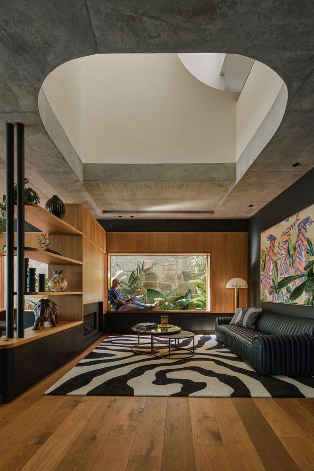 A sophisticated living room with warm timber wall panelling and built-in shelving, a dark ribbed sofa, sculptural coffee tables, and a bold black-and-cream abstract rug. The room features a dramatic concrete ceiling with a large rounded recessed void or skylight, dark painted walls, a large artwork, and a picture window overlooking greenery and a stone wall.