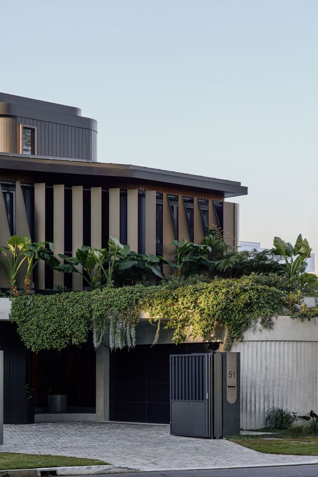 A contemporary multi-level residential facade with a flat roof and rounded upper-storey form. The dark charcoal facade combines vertical ribbed cladding, slim windows, projecting roof planes, and contrasting pale vertical fins. Dense cascading greenery softens the front terrace and boundary wall, with a dark pedestrian gate, driveway, and house number visible at street level.