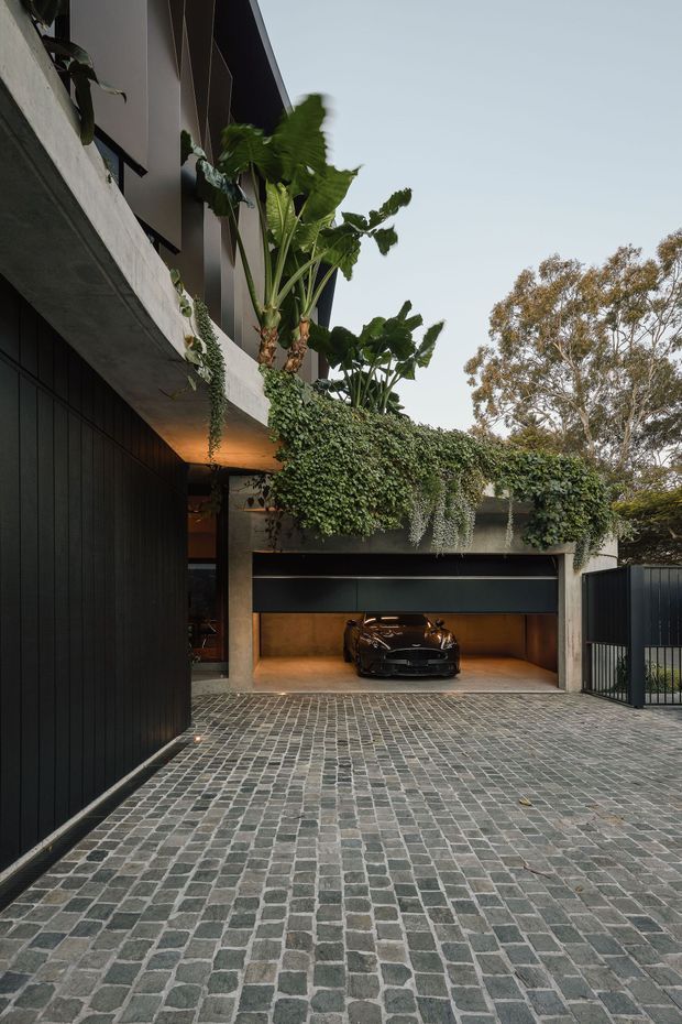 A contemporary residential exterior viewed from the driveway, featuring a dark vertical-clad facade, exposed concrete balcony and garage structure, lush cascading planter vegetation, and a single-car garage with a luxury vehicle inside. The foreground is a broad driveway paved in small grey cobblestones, with dark metal fencing and mature trees around the property.
