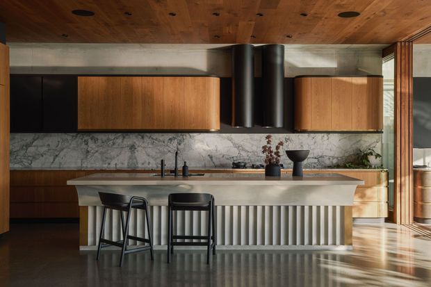 A refined contemporary kitchen with a large sculptural island, fluted vertical detailing, dark stone flooring, marble-look splashback, and warm timber cabinetry. The ceiling is clad in timber, with dark cylindrical rangehood elements and expansive glazing or doors at the right bringing in strong natural light. Two black bar stools sit at the island, creating a sophisticated, warm, high-contrast interior.