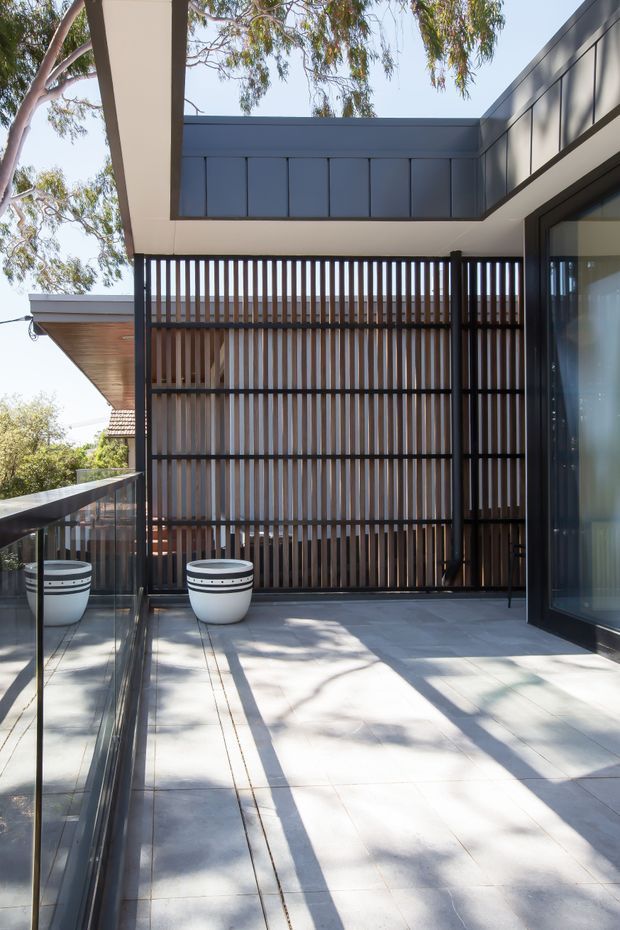 A bright modern outdoor terrace or balcony with large pale grey tiled flooring, a dark metal-framed glass balustrade, and a striking vertical timber privacy screen. The terrace is enclosed by contemporary flat-roofed architectural forms with dark grey cladding, recessed glazing, and a warm timber soffit. Two white cylindrical planters sit against the screen, while surrounding eucalyptus branches and neighbouring roof forms are visible beyond.