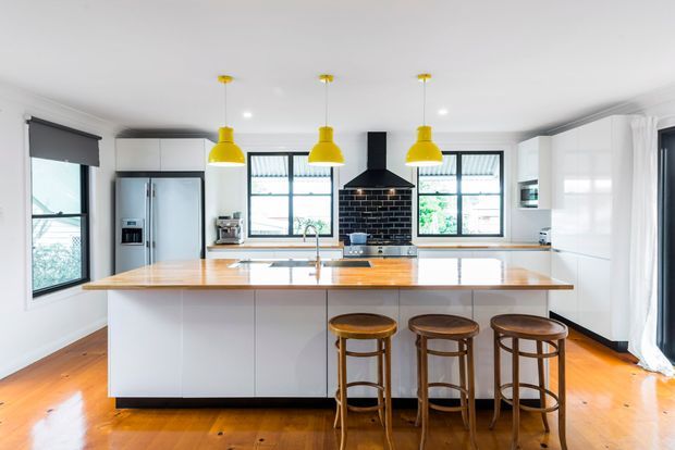Bright contemporary kitchen with a large white island, timber benchtop, integrated sink, three timber bar stools, white cabinetry, stainless-steel appliances, black brick splashback and rangehood, yellow pendant lights, and warm timber flooring. Large windows and white curtains provide abundant natural light.