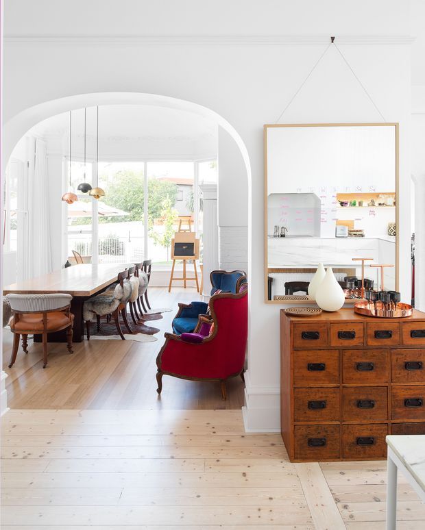 Bright, refined residential interior showing a white hallway or entry space opening through a large arched opening into a dining area. The dining room contains a long timber table with upholstered and timber chairs, pendant lights, and large glazed doors overlooking greenery. A vivid red and blue upholstered armchair sits in the foreground beside a vintage timber drawer chest and large framed artwork or mirror. A kitchen or work area is visible through the framed opening on the right. The space combines white painted architectural detailing, pale timber flooring, traditional furniture, and contemporary brightness.