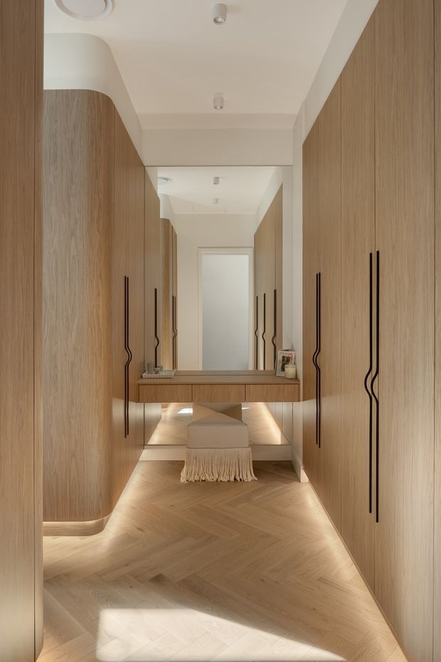 A refined dressing corridor or walk-in wardrobe lined with full-height, light timber cabinetry. Curved cabinet ends, elongated black sculptural handles, a central floating console with integrated lighting, and a large mirror create a warm, luxurious composition. The space has a pale herringbone timber floor, soft off-white walls and ceiling, concealed under-cabinet illumination, and a small upholstered bench with a fringed throw.