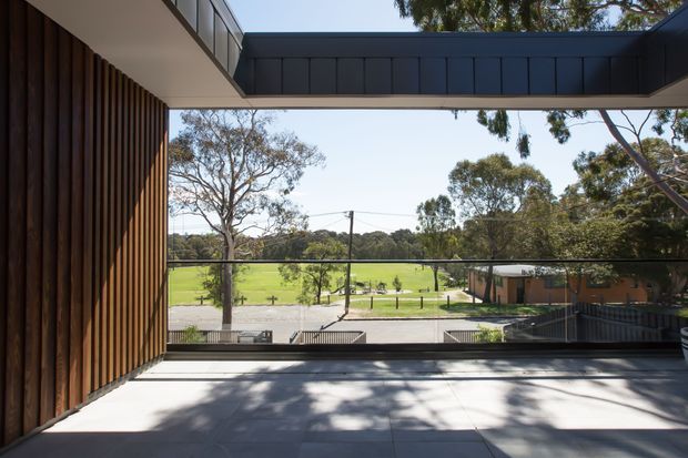 A contemporary covered terrace or balcony overlooking a green open field and mature trees. The space features large-format grey paving, slim dark metal balustrades, a dark projecting roof or bridge element, and a vertical timber batten wall on the left. The composition frames the landscaped outlook through an open glazed or balustraded edge.