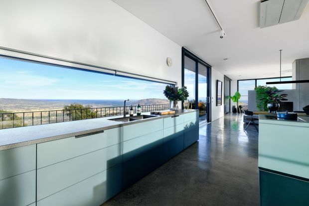 A bright, contemporary open-plan kitchen with a long pale built-in island and extensive floor-to-ceiling glazing. The kitchen opens toward a balcony with expansive landscape views, while polished concrete flooring, dark-framed windows, streamlined cabinetry, and integrated appliances create a clean modern interior. A dining and living area is visible beyond the kitchen.