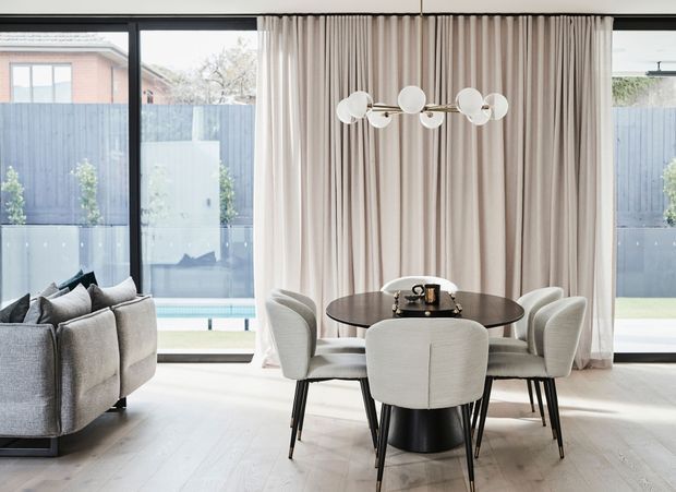 Bright, refined open-plan dining and living area with a round dark dining table, upholstered dining chairs, sculptural globe pendant light, soft full-height curtains, pale timber flooring, and expansive black-framed glazing opening toward a private courtyard and pool. A contemporary grey sofa is partially visible at left.