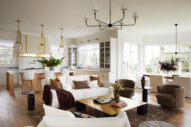 Bright open-plan interior combining a white kitchen, living room, and dining area. The space features extensive white-framed glazing, light timber flooring, pale cabinetry, brass pendant lights, a large cream sectional sofa, patterned rug, and warm wood and brass accents. Built-in glass-front cabinetry and a central black-and-brass chandelier add a refined traditional character.