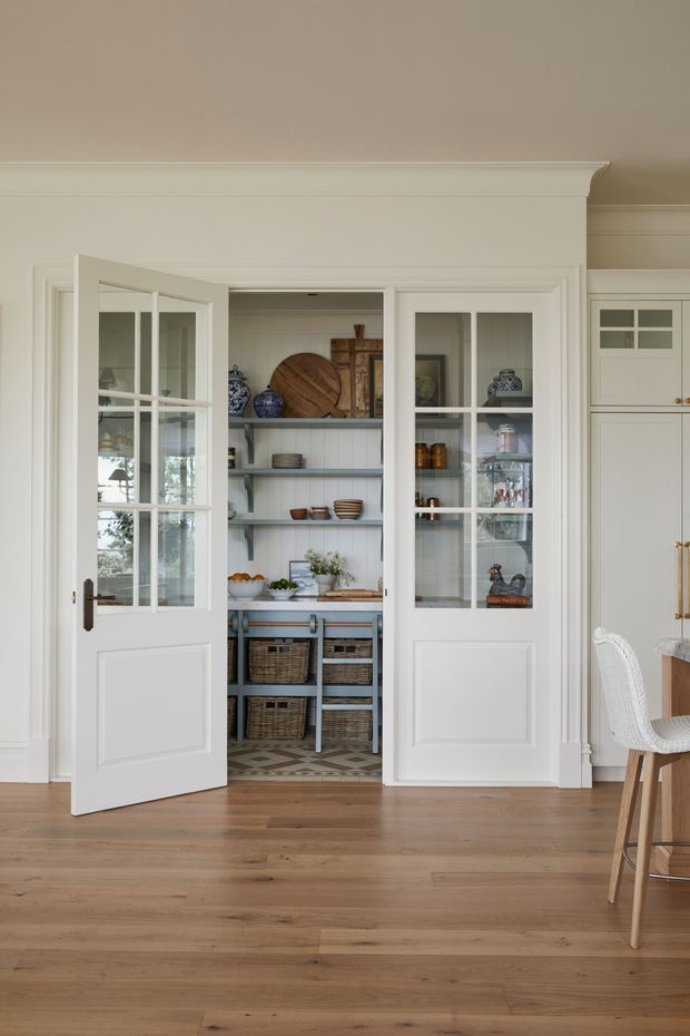 A bright, refined interior view of a built-in walk-in pantry behind open white French doors. The pantry features open shelving, white vertical wall panelling, baskets, jars, crockery and timber accents, with a patterned tiled floor. The foreground has warm timber flooring, white panelled walls and detailed traditional trim; part of an adjacent dining area with a white woven chair and timber cabinetry is visible at right.