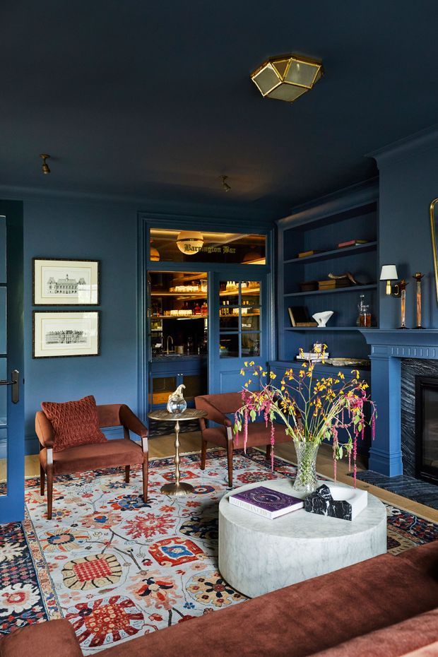 A richly layered, dark blue living room with built-in shelving, a fireplace surround, framed artwork, and a mirrored or glazed bar cabinet. Rust-coloured upholstered armchairs and sofa surround a rounded pale stone coffee table, while a colourful patterned rug and fresh floral arrangement add vivid contrast. Decorative brass lighting and traditional wall mouldings contribute to a refined, eclectic atmosphere.
