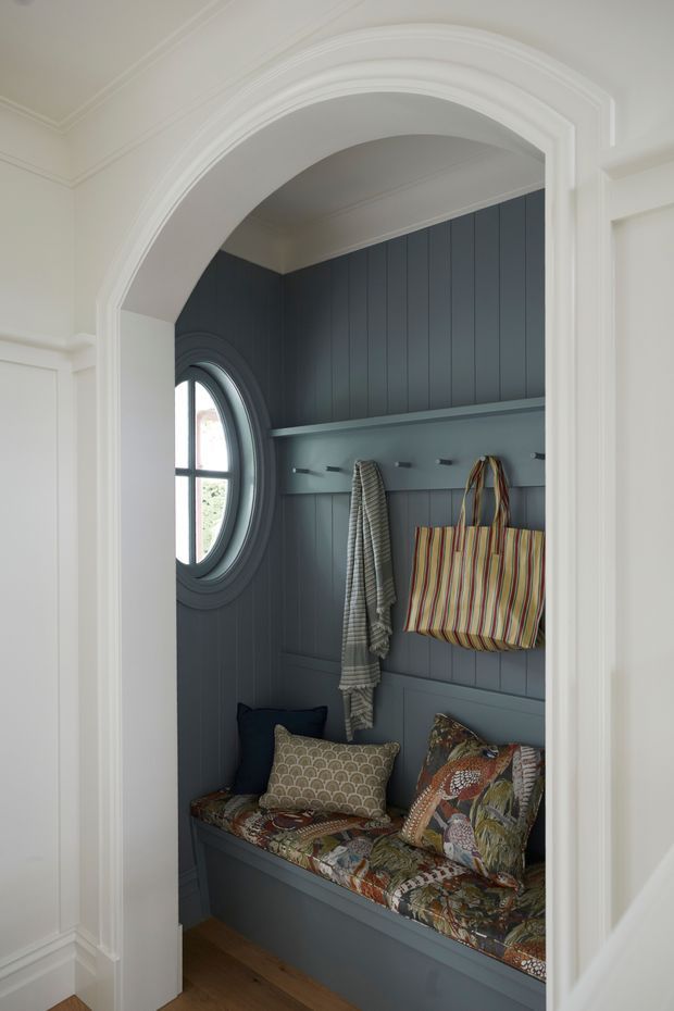 A compact, built-in entry nook viewed through a white, classically moulded arched doorway. The nook features dark blue-grey vertical wall panelling, a rounded window with deep trim, a long shelf with hooks, a built-in bench with patterned cushions, and hanging bags and textiles. White decorative mouldings and warm timber flooring frame the space.