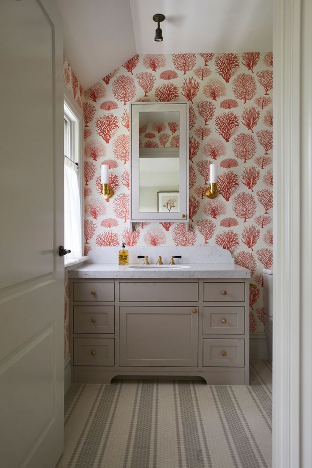 A compact, traditionally styled bathroom viewed through an open doorway. The room features a taupe-gray freestanding vanity with raised-panel cabinetry and brass knobs, a pale stone countertop with a centered basin and brass faucet, a tall framed mirror, matching brass wall sconces, coral-red botanical wallpaper, and a patterned gray-and-white tiled floor. A window provides natural light from the left.