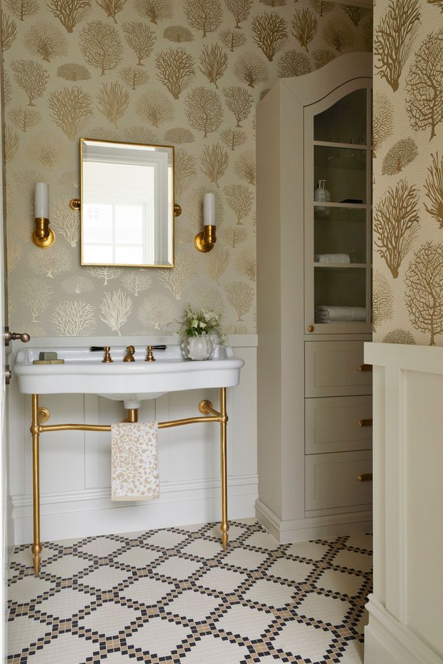 A refined traditional bathroom with a wall-mounted white porcelain basin, exposed brass plumbing and towel rail, brass wall sconces, framed mirror, patterned botanical wallpaper, built-in cabinetry with arched display shelving, and a decorative geometric mosaic tile floor. The palette combines warm white, taupe, muted botanical tones, and polished brass.
