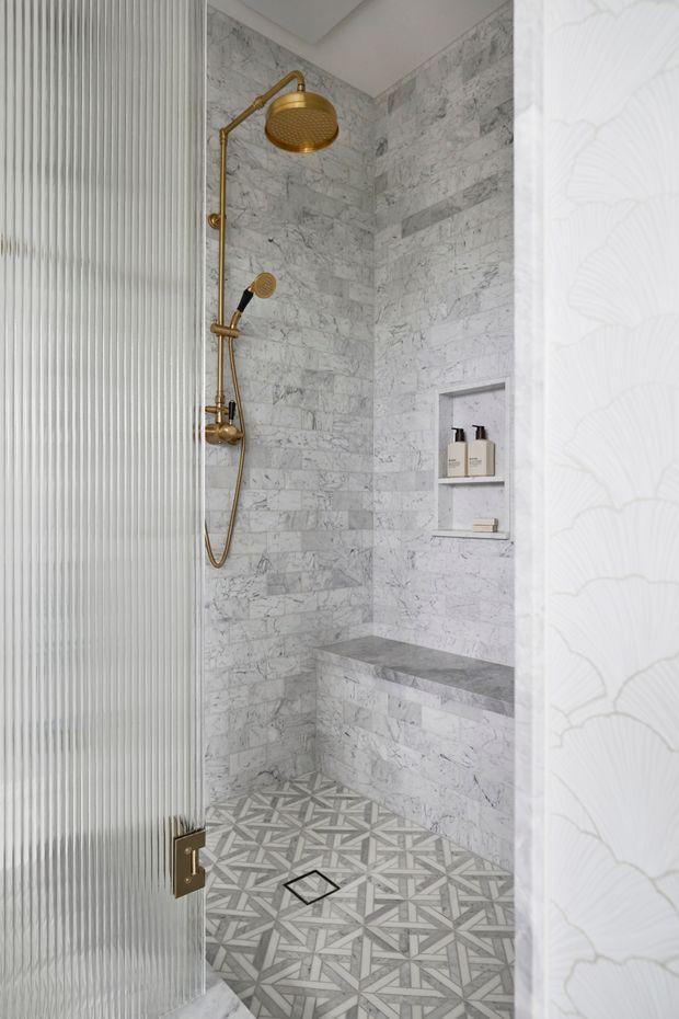 A compact, refined walk-in shower with marble-look wall tiles, a geometric patterned floor, brushed gold shower fittings, a built-in recessed niche, and a stone bench. The shower is partially enclosed by a fluted glass door, with subtle patterned wallcovering visible at the entrance.