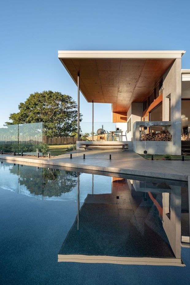 Exterior view of a contemporary residential home beside a still reflecting swimming pool. The house features a broad flat roof with deep overhanging eaves, a warm timber-lined soffit, rendered walls, extensive glazing, slender dark supports, and an integrated outdoor fireplace or wood-storage feature. A glass pool fence and landscaped garden are visible.
