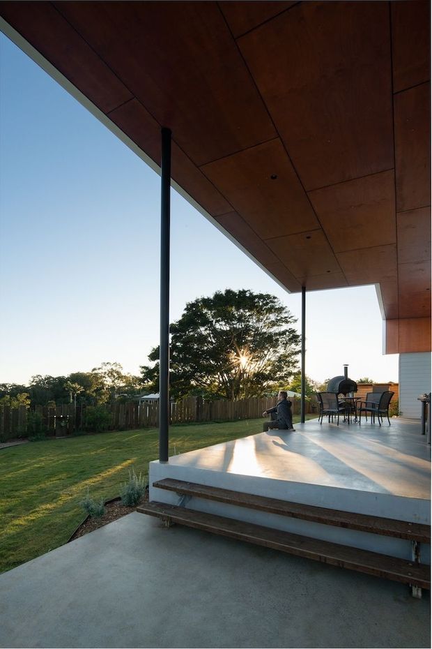 A covered outdoor terrace viewed from the garden, featuring a broad concrete deck, timber-clad soffit, slender dark structural posts, and wide steps leading up from a lower concrete surface. Outdoor dining furniture and a barbecue sit at the far end, with a fenced lawn and mature tree beyond. Warm late-afternoon sunlight creates long shadows across the terrace.