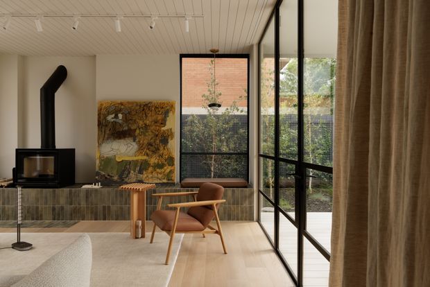 A refined contemporary living room with a black freestanding wood stove, textured tiled hearth wall, timber flooring, a sculptural lounge chair, and a large abstract artwork. Full-height black-framed glazing opens toward a planted courtyard, while linen curtains and a slatted white ceiling add warmth and texture.