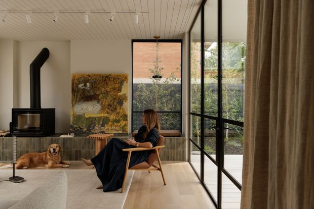 Warm, refined living room with a black wood-burning stove set against a textured tiled feature wall, a low light-wood media cabinet, and an abstract artwork. A woman sits in a timber-framed lounge chair beside a large black-framed glazed wall and garden courtyard. The room has pale timber flooring, a light textured rug, a slatted white ceiling with track lighting, and full-height neutral curtains.