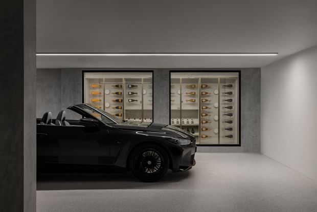 A dark luxury sports car is displayed in a minimalist enclosed garage or showroom. The space has smooth grey concrete-like walls, a pale seamless floor, recessed linear ceiling lighting, and two illuminated glass-fronted display cabinets containing wine bottles and glassware. The composition is moody and highly refined, with strong contrast between the black vehicle and neutral architectural finishes.