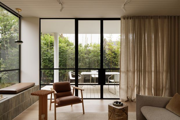 A refined contemporary living room with full-height black-framed glazing opening to a small deck and lush garden. The room features a light timber slatted ceiling, built-in timber cabinetry with a window seat, floor-to-ceiling linen curtains, a brown leather lounge chair, and a neutral upholstered sofa. A sculptural pendant, textured timber side table, and warm natural materials create a calm, understated interior.