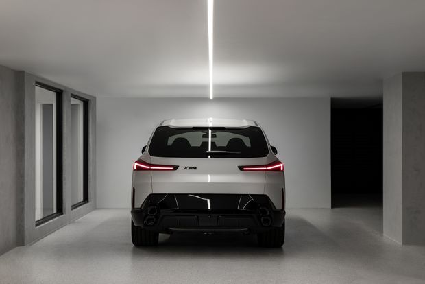A minimalist enclosed residential-style garage with a white SUV parked centrally, viewed from the rear. The space has smooth grey concrete-like walls and floor, dark-framed windows along the left wall, a dark open doorway at the rear right, and a single linear ceiling light. The composition is monochromatic, clean, and highly contemporary.