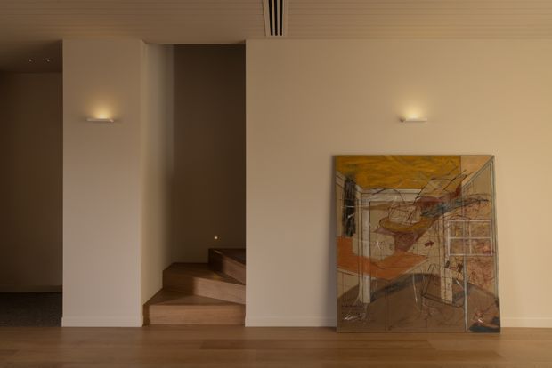 A refined, warmly lit interior hallway or landing with a minimalist stairwell. The space features smooth off-white walls, integrated wall sconces, a timber floor, a timber-toned stair, a linear ceiling air vent, and a large abstract artwork leaning against the wall.