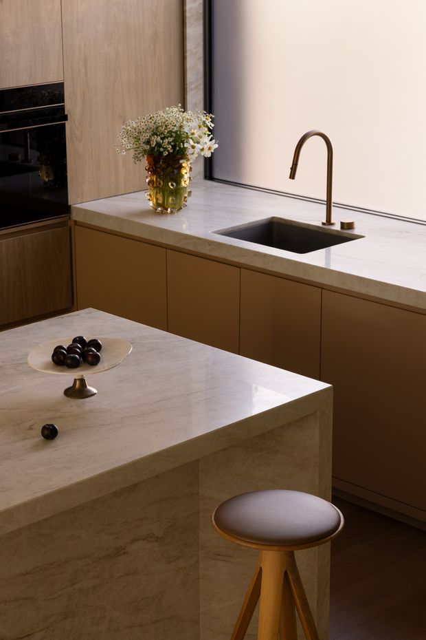 A refined contemporary kitchen featuring a large pale veined stone island, matching stone benchtops, warm wood-toned cabinetry, integrated dark appliances, and a deep undermount sink with a bronze gooseneck tap. A timber stool and understated floral arrangement add warmth to the sophisticated, softly lit space.