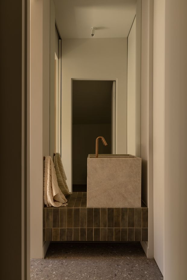 A narrow, warmly lit bathroom or powder room viewed through a doorway. The space features a wall-mounted vanity clad in dark vertically arranged tiles, a large rectangular stone or plaster-finished vessel basin, and a curved brass tap. A full-height mirror reflects the compact room, while folded towels rest on the vanity. The floor is finished in a speckled terrazzo-like material, and the overall design is minimalist and refined with earthy neutral tones.