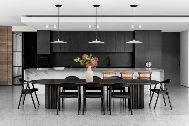 Contemporary open-plan kitchen and dining area with a dark timber or charcoal kitchen wall, black cabinetry, integrated appliances, a dark stone-look splashback, and a pale veined stone island. A long dark dining table with black chairs sits in front of the island, with three stools along the counter. Minimal pendant lighting, pale terrazzo-like flooring, and a restrained monochrome palette create a refined, modern atmosphere.