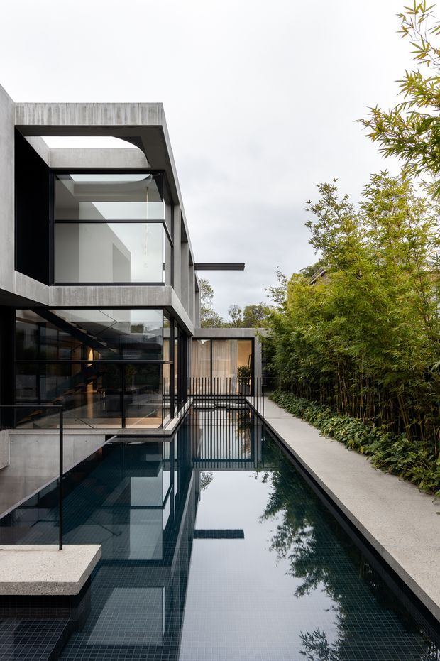 Exterior view of a contemporary multi-level residence with exposed concrete framing, extensive black-framed glazing, a narrow reflecting or swimming pool alongside the building, and a landscaped bamboo-lined walkway. The architecture features strong rectilinear forms and a projecting roof or canopy.