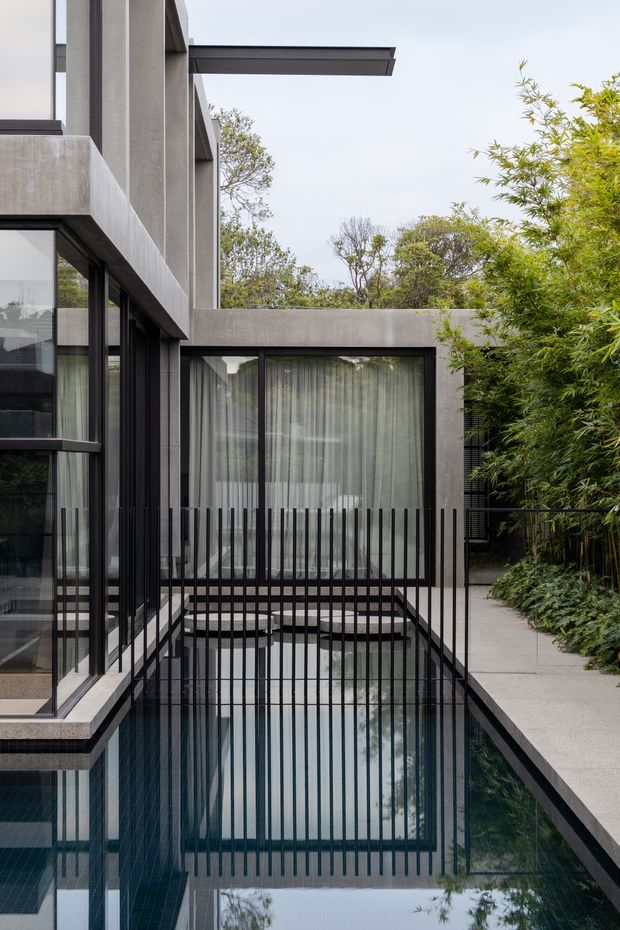 A contemporary courtyard exterior focused on a narrow reflecting pool beside a dark-framed glazed residence. The home has monolithic grey concrete walls and roof forms, extensive black-framed glazing, sheer curtains, and a black vertical-bar balustrade spanning the pool edge. A pale concrete walkway and dense bamboo-like planting border the right side, creating a private, minimalist courtyard atmosphere.