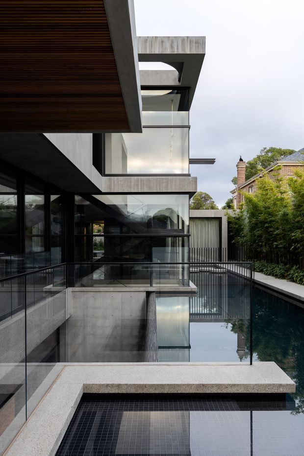 A contemporary multi-level residential exterior viewed from an elevated terrace or balcony. The building features strong rectilinear concrete forms, extensive dark-framed glazing, projecting slabs, a timber-lined soffit, glass balustrades, and black metal railings. A reflective tiled pool occupies the foreground and continues alongside the building, with dense planting and a neighbouring traditional brick house visible beyond. The composition emphasises layered indoor-outdoor connections, crisp geometry, and contrasting concrete, glass, timber, and dark metal.