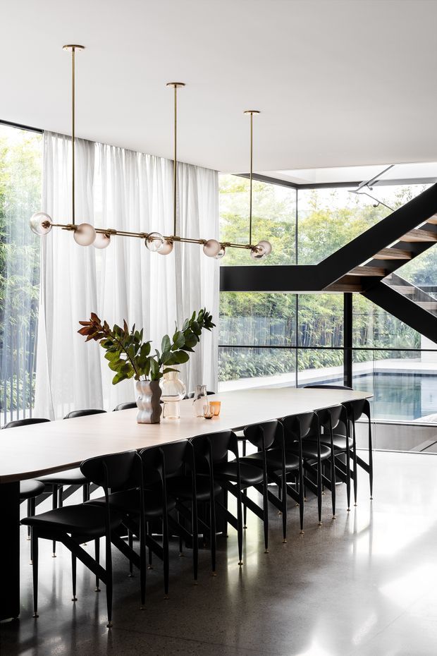 A spacious, light-filled contemporary dining area with a long white dining table surrounded by black chairs. Floor-to-ceiling glazing and sheer curtains overlook lush greenery and an indoor pool, while a dramatic black open-riser staircase crosses the background. A linear brass-and-glass pendant light hangs above the table, with a simple floral arrangement as the centerpiece.