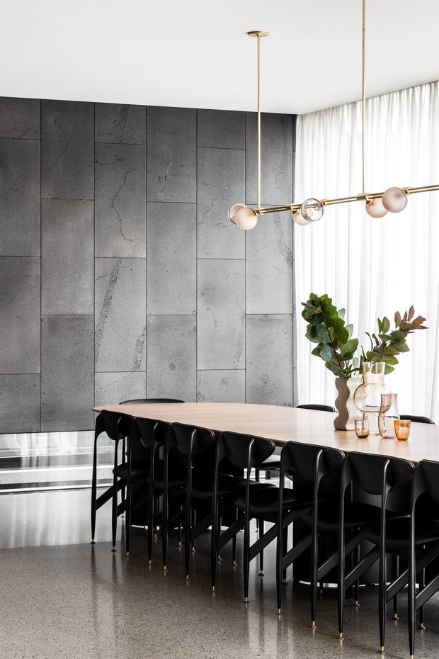 Contemporary dining area with a long light-toned dining table and numerous black dining chairs, set against a dramatic wall of large-format grey stone-look panels. A linear brass pendant with spherical glass lights hangs above the table, while sheer full-height curtains provide soft daylight. The polished grey floor reflects the furniture and lighting, creating a refined, high-contrast interior.