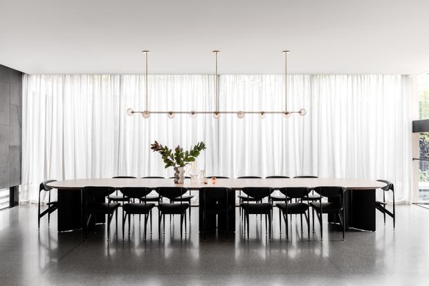 A spacious contemporary dining room with a long pale stone or solid-surface dining table surrounded by black dining chairs. Full-height sheer white curtains provide abundant natural light, while a linear pendant with small spherical lights hangs above the table. The room has dark built-in cabinetry or wall elements, a polished grey floor, and a restrained monochromatic palette.