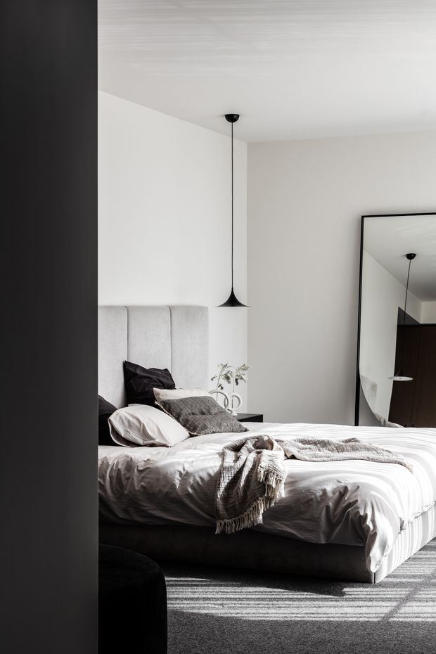 A calm, refined contemporary bedroom viewed through a dark doorway. The room features a large upholstered bed with a tall vertically panelled light-grey headboard, white and grey layered bedding, a slim black pendant light, a small dark bedside table, and a large black-framed leaning mirror reflecting the pendant and dark timber joinery. The palette is predominantly white, light grey, charcoal, and black, with soft natural light and textured carpet.