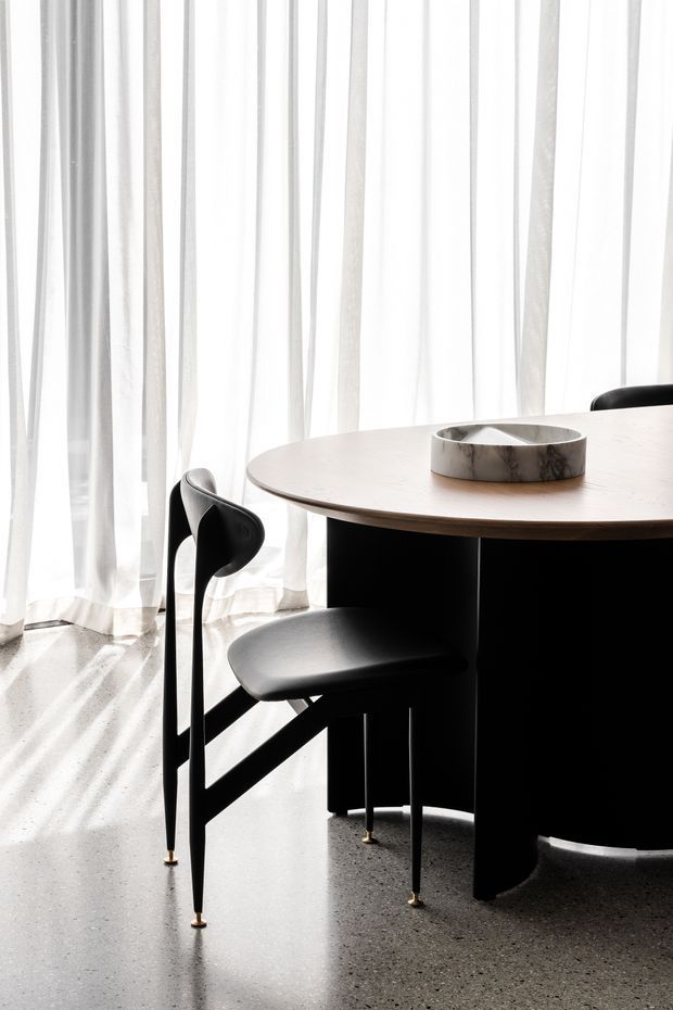 A minimalist dining area with a round light timber dining table, sculptural black dining chairs, and a dark central pedestal base. Sheer white curtains diffuse strong daylight across a polished grey floor, while a shallow stone or marble bowl sits at the centre of the table.